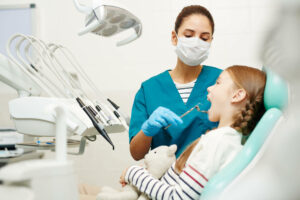 Serious concentrated young female dentist in mask and gloves examining childrens teeth in dental room, redhead girl with toy keeping mouth open at checkup